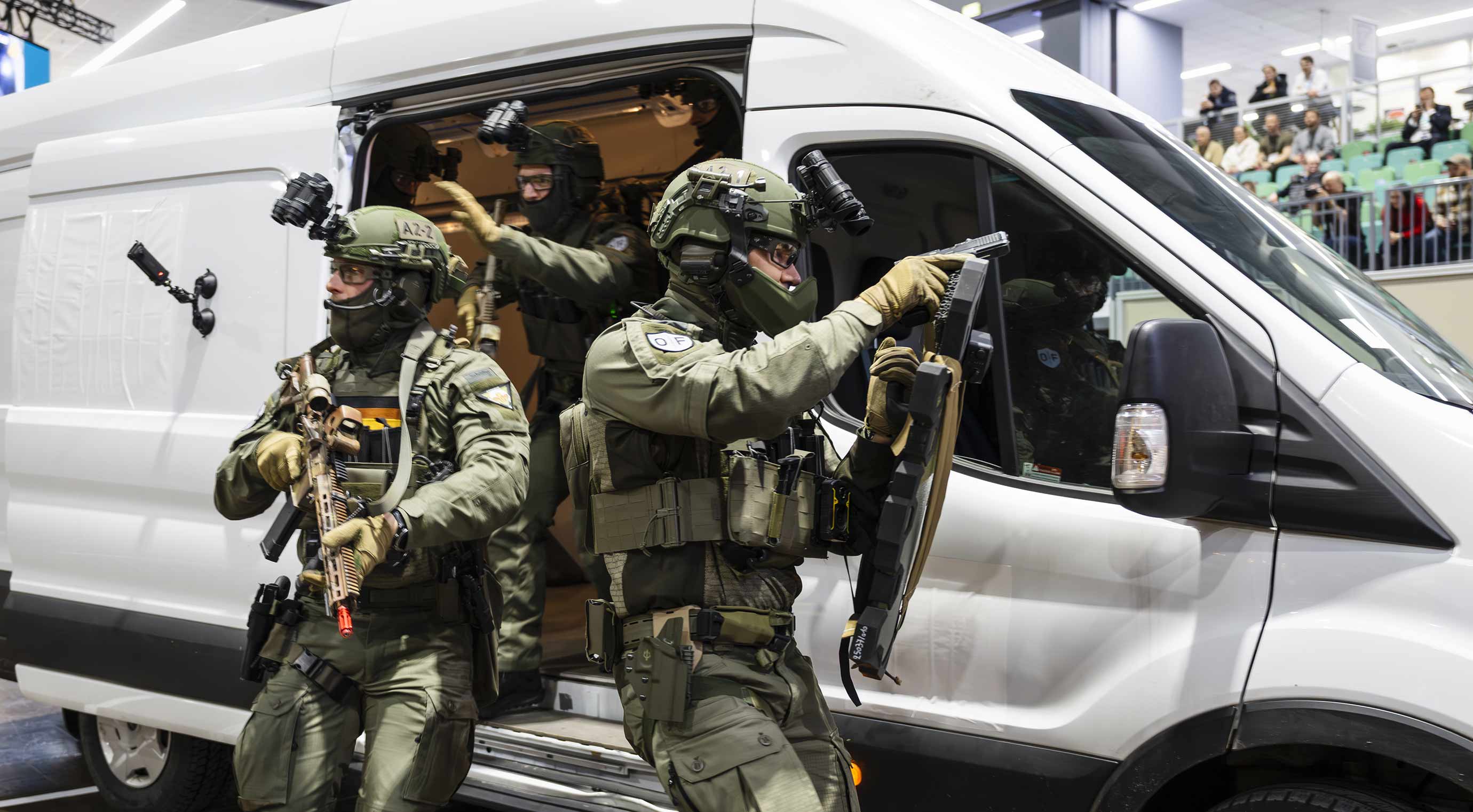 Special forces unit steps out of a van during a demonstration, carrying shields, helmets, and weapons, in an exhibition hall.