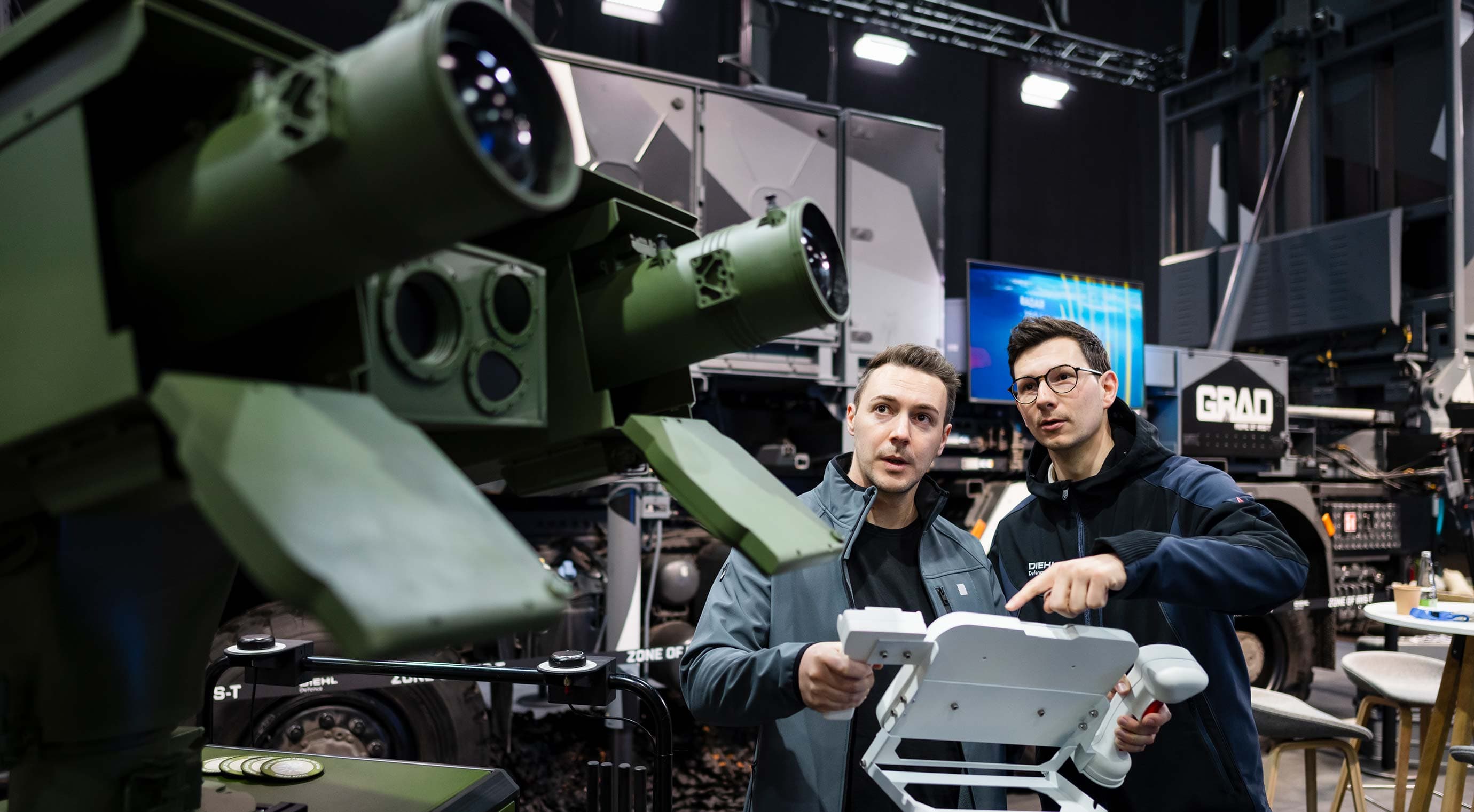 Product presentation of a military sensor unit; two people are operating the control unit at the stand in the exhibition hall.
