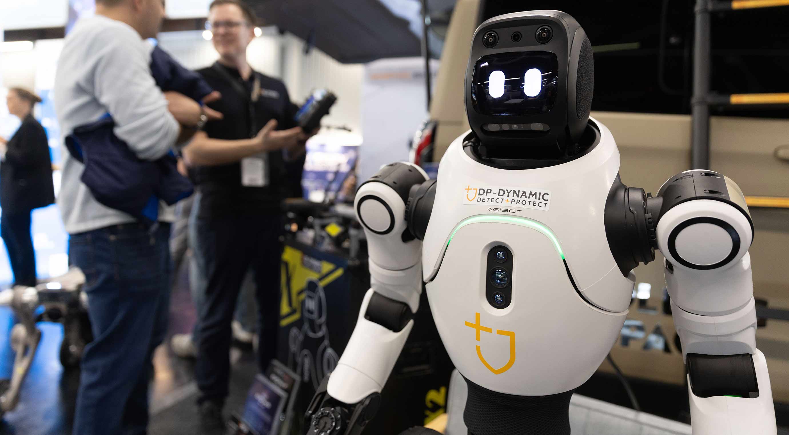 Humanoid security robot at a trade fair stand, with a display face and sensors.